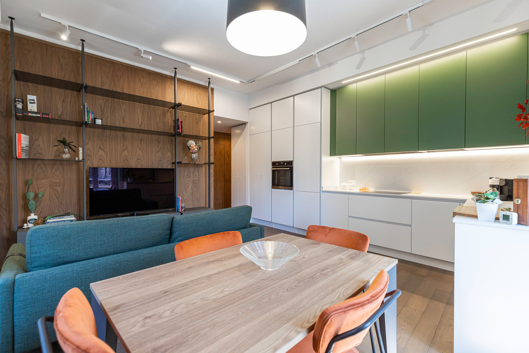 Zona living_libreria Una sala con libreria marrone a parete, divano e tavolo da pranzo. La spaziosa cucina bianca e verde, luminosa e funzionale, completa l’ambiente